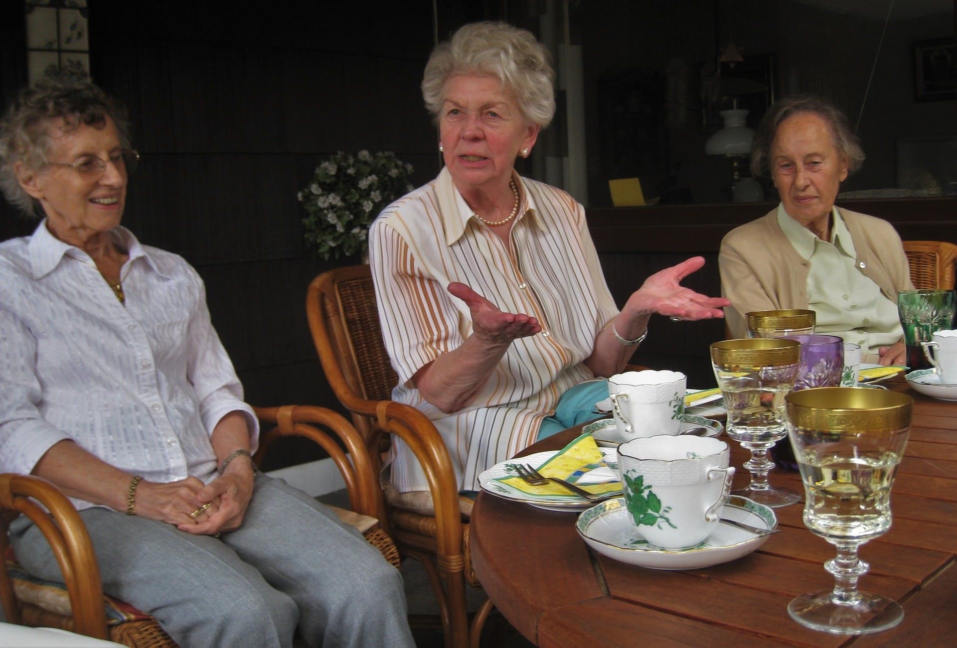 Three older adults seated at a dining table, talking over tea and glasses in a cozy indoor setting