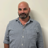 Man standing indoors against a beige wall, wearing a blue and white checkered shirt, with a slight smile.