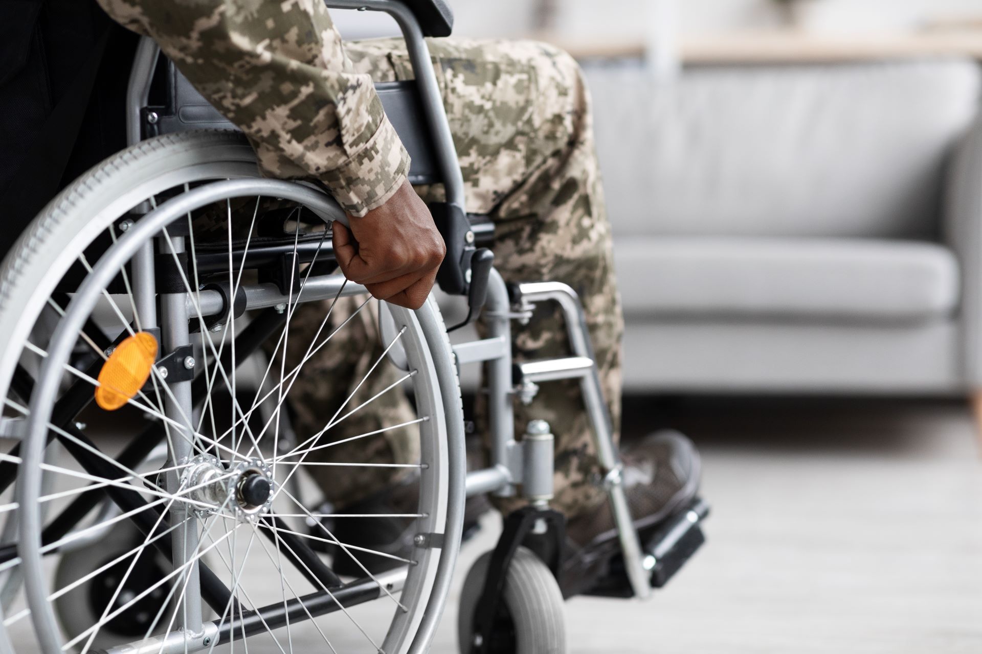 Person in camouflage clothing pushing a wheelchair indoors near a gray sofa
