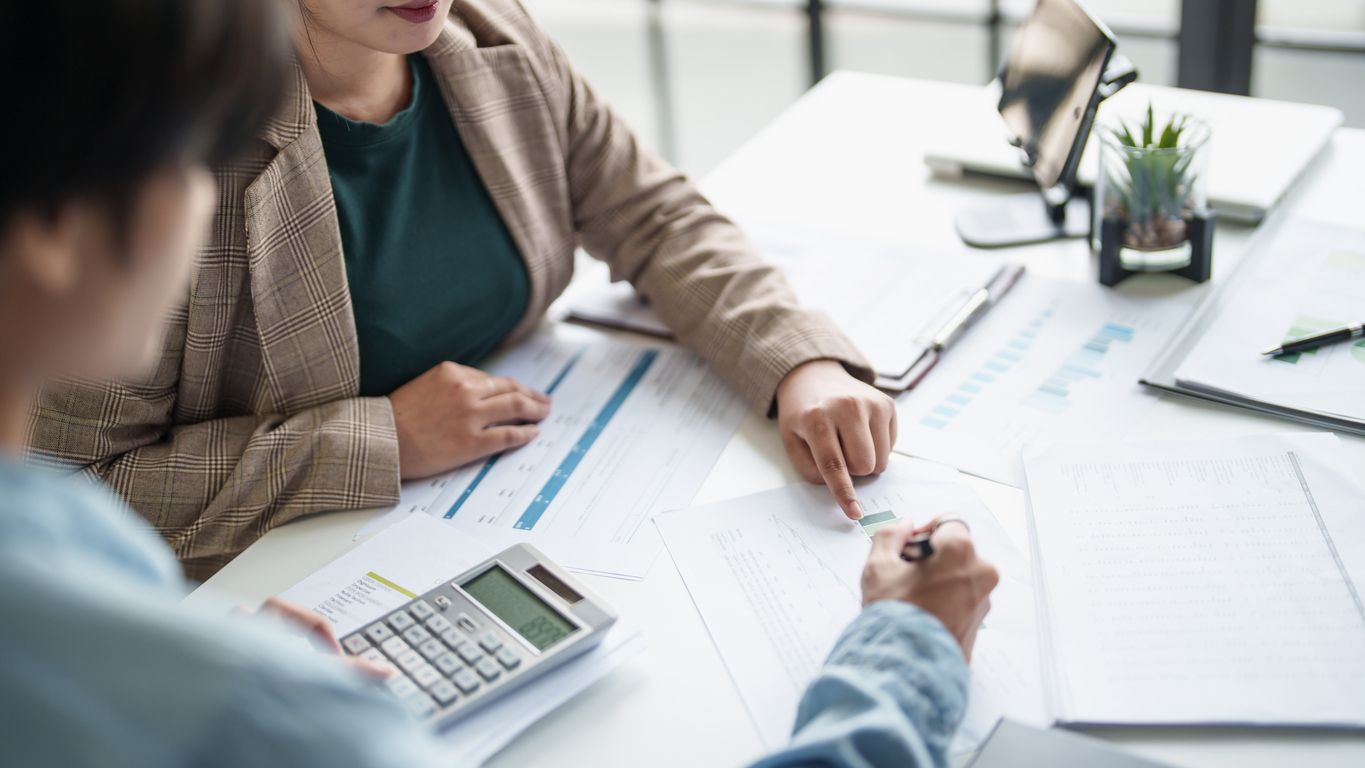 Two people reviewing paperwork at a desk, one points, another uses a calculator.