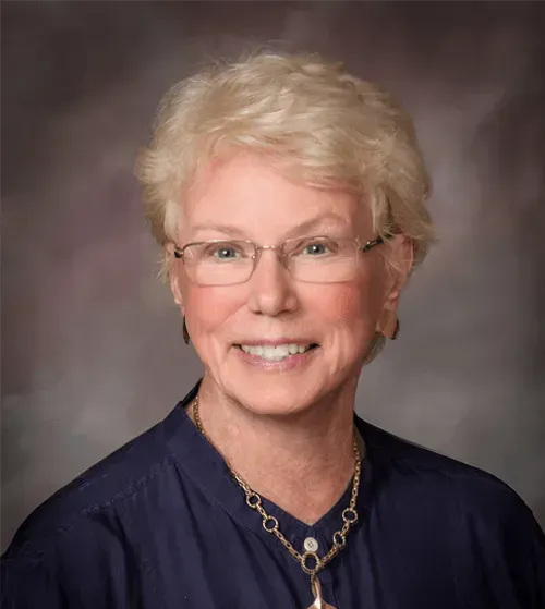Woman with short blonde hair and glasses smiles. Wearing a blue top and a necklace; background is blurred.
