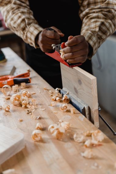 Hands planing a wooden board with a red hand plane, wood shavings scattered on the workbench.