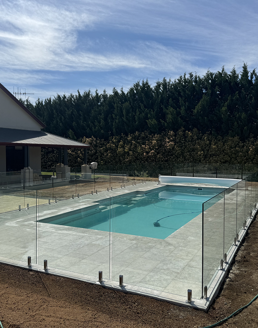 Swimming pool surrounded by glass fencing on a sunny day. Green hedge in the background.