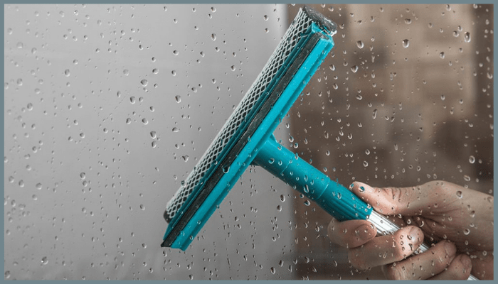 Hand using a squeegee to clean water droplets from a glass pool fence panel.