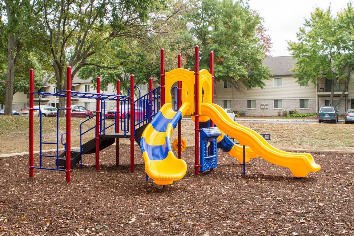 Playground with yellow and blue slides, climbing structures, and red support beams, on a wood chip base.