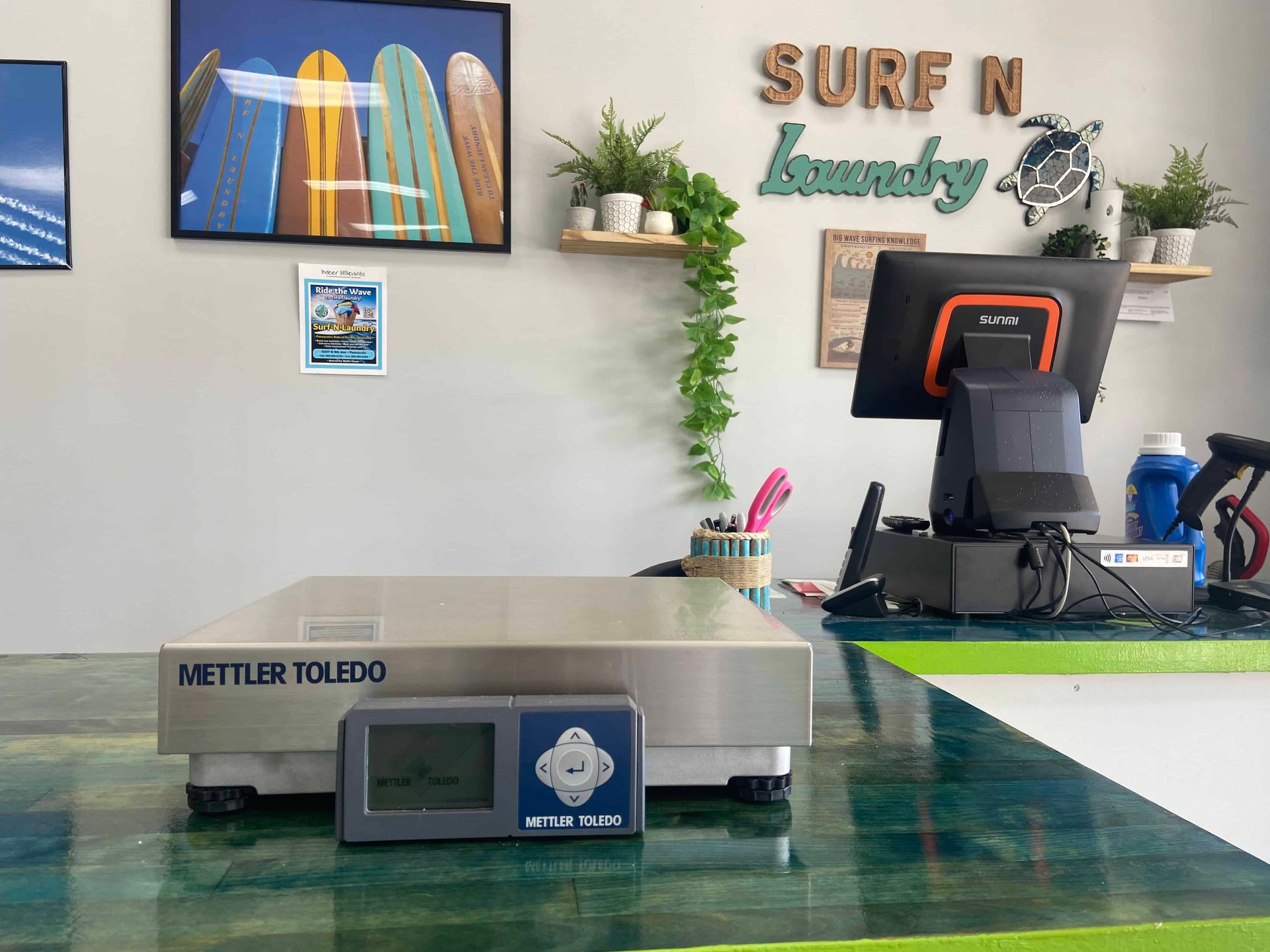 Our scale that we use to weigh dirty laundry coming in to be cleaned through our Wash and Fold service with our logo of Surf -N- Laundry in the background with a Payment terminal in front