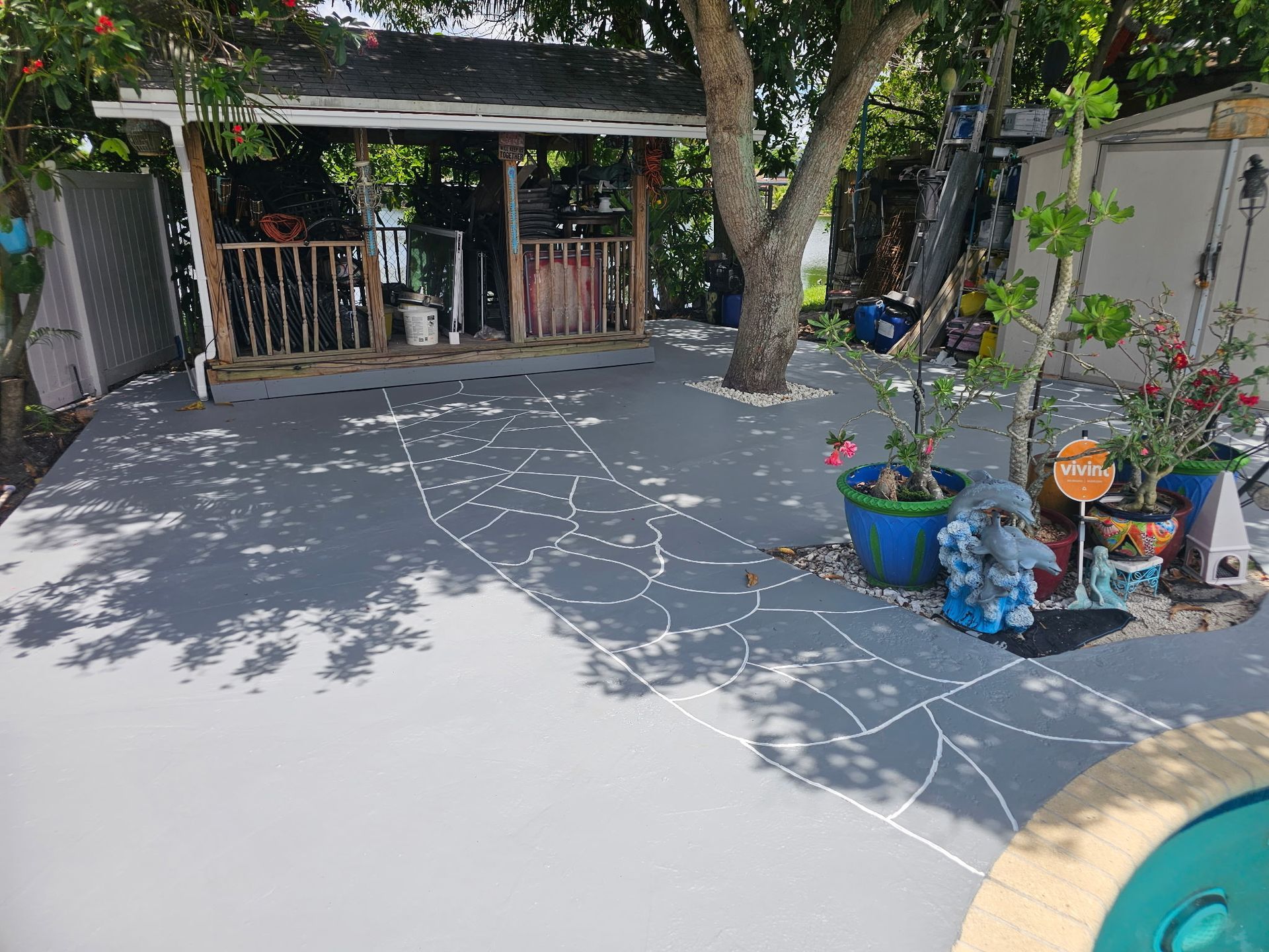 Sunlit concrete patio with a tree, potted flowers, and a shaded porch in the background