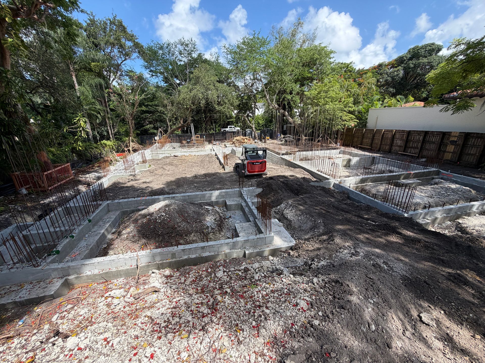 Construction site with excavated foundation, concrete forms, and a small red skid steer under trees