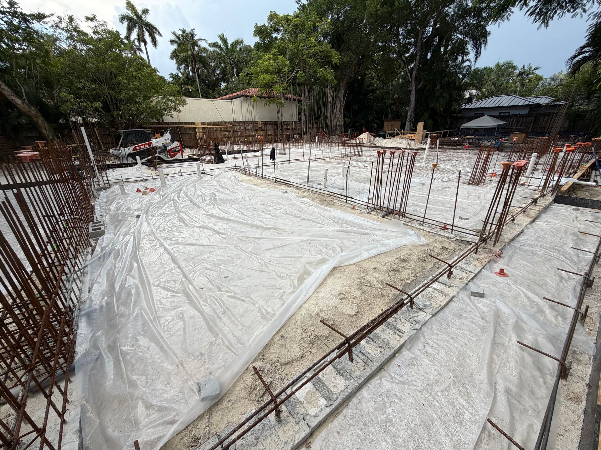 Construction site with concrete foundation covered by white tarp and exposed rebar under trees