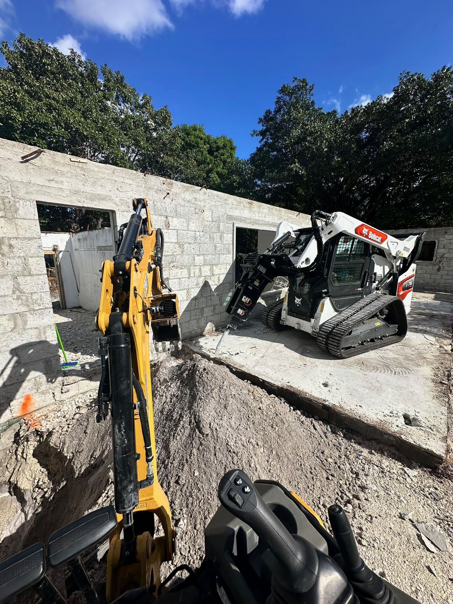 Excavator demolishes a concrete wall beside a damaged skid steer under a blue sky.
