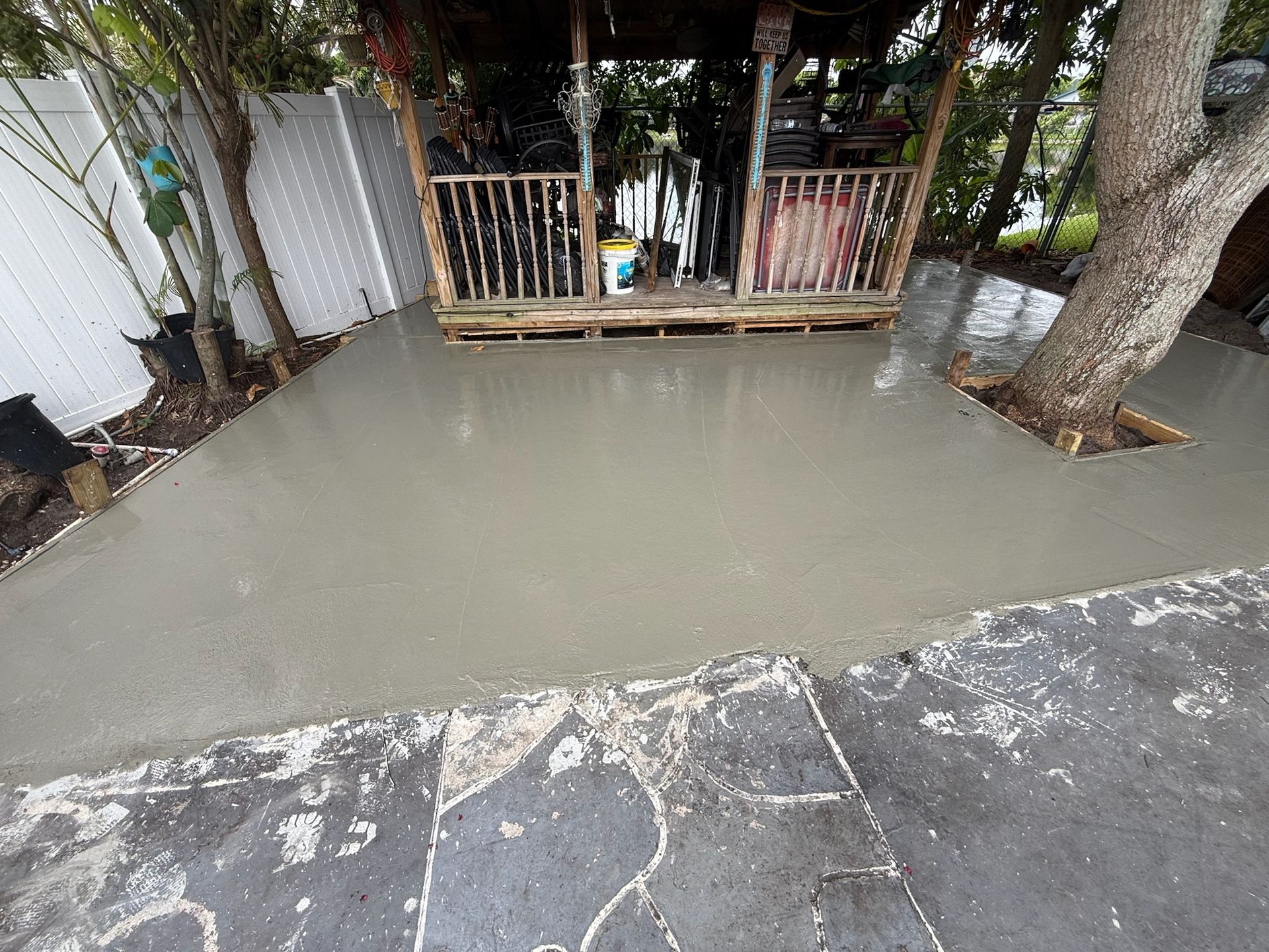 Flooded concrete yard beside a porch and tree, with muddy water pooled across the ground.