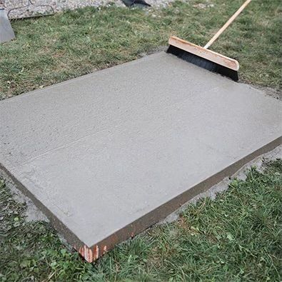 Freshly smoothed concrete slab outdoors with a broom on top, surrounded by grass