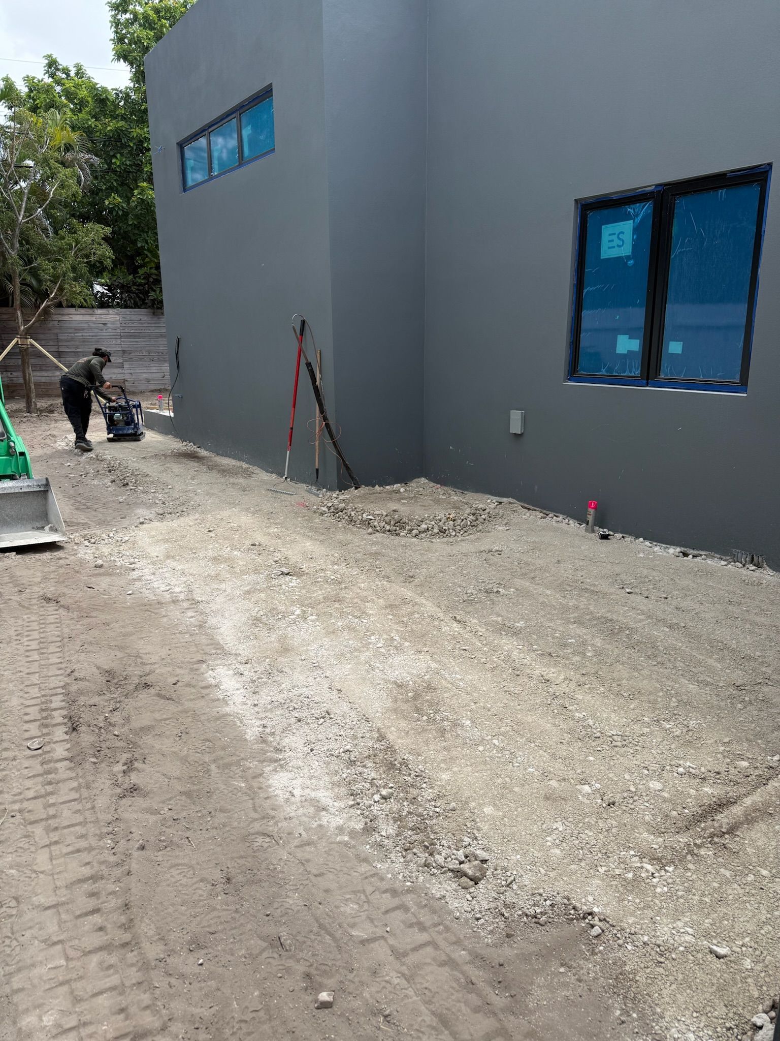 Gray building with blue windows beside a gravel path under construction, with workers and tools nearby
