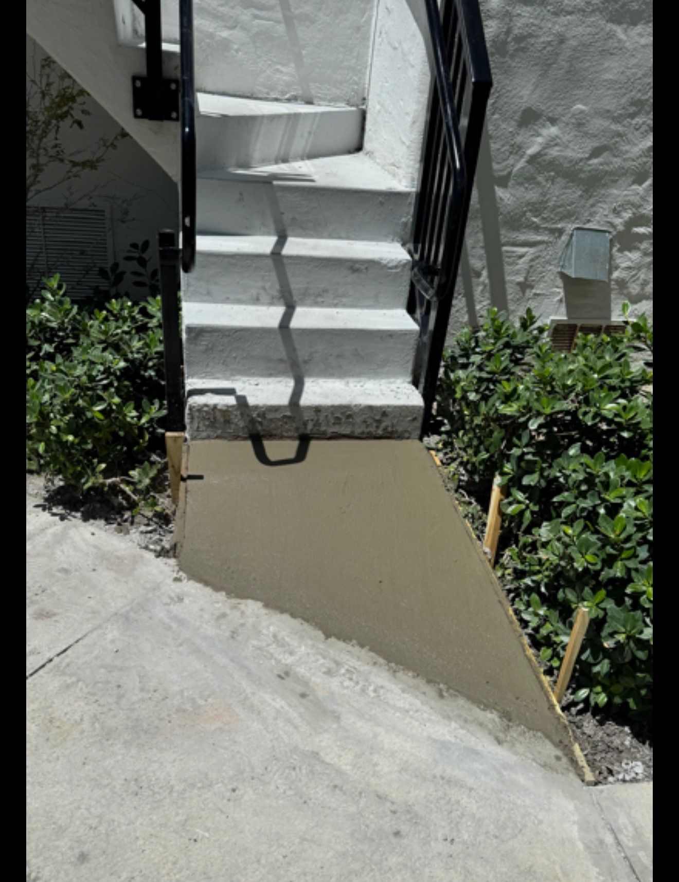 Concrete outdoor staircase with white steps and black railing beside a sloped wall and plants