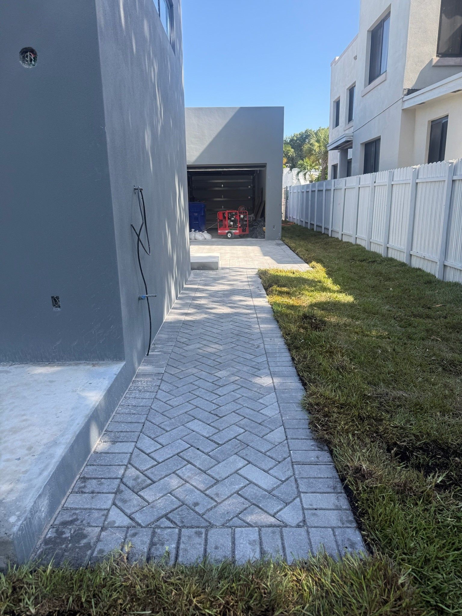 Brick walkway beside a gray house and white fence, leading to a garage entrance with a narrow grass strip.