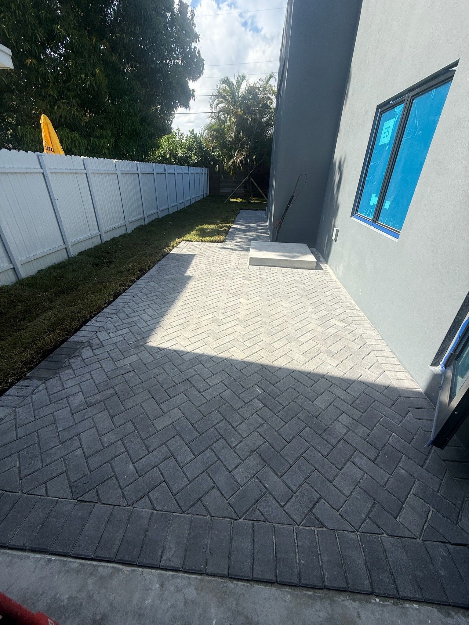 Paved side yard beside gray house with blue window, white fence, and trees in sunlight