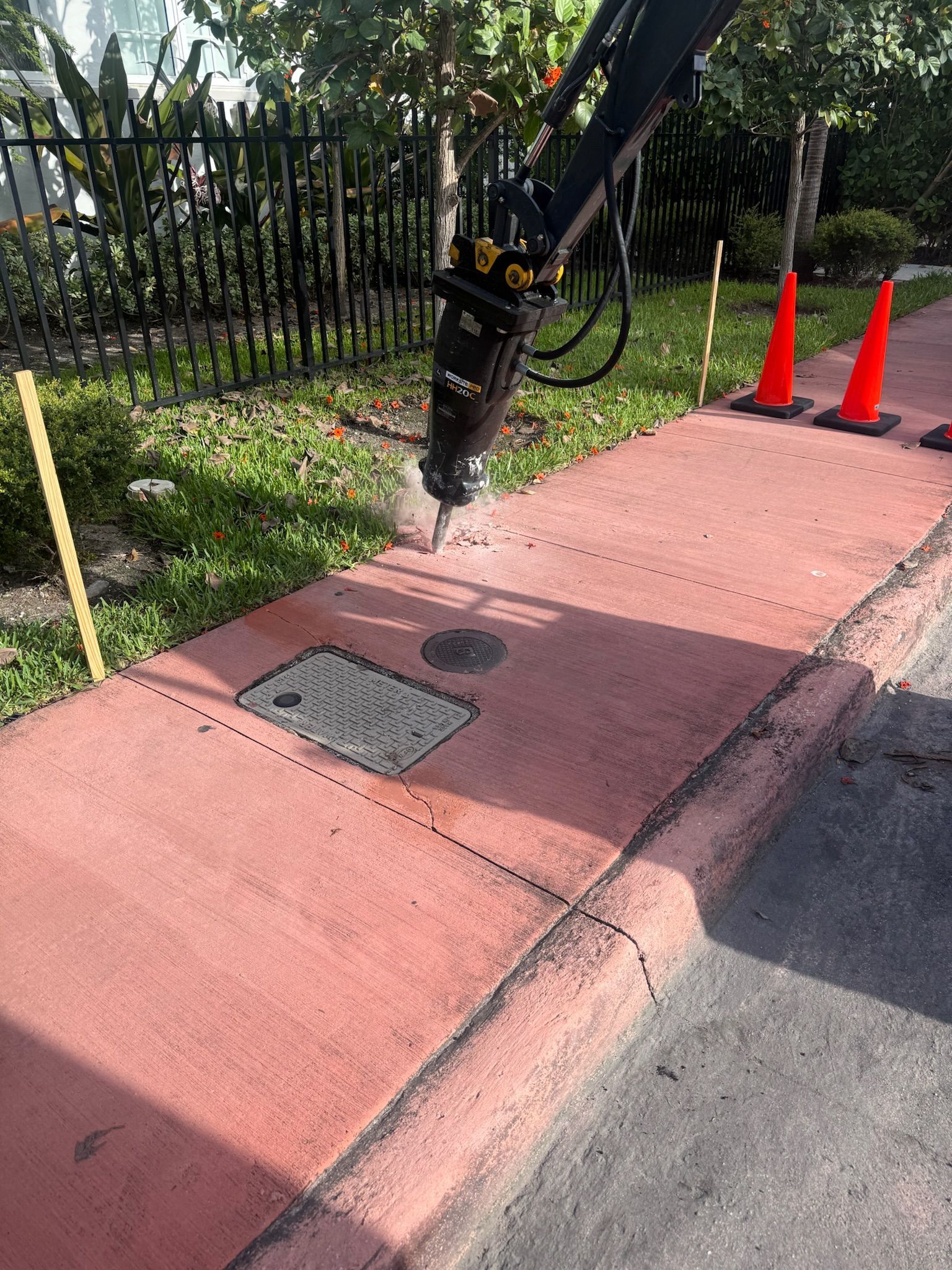 Roadside crew jackhammering a red sidewalk near a black fence, with orange cones marking the area