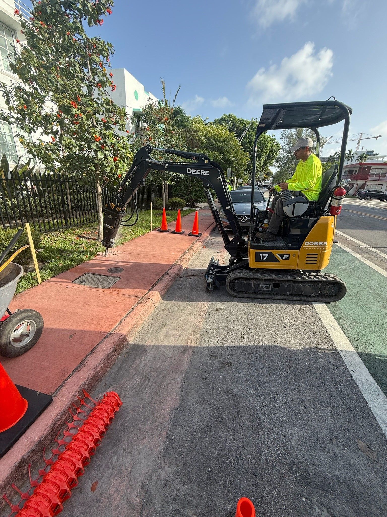 Small excavator on a paved path beside orange cones and red mats near trees in a park-like area