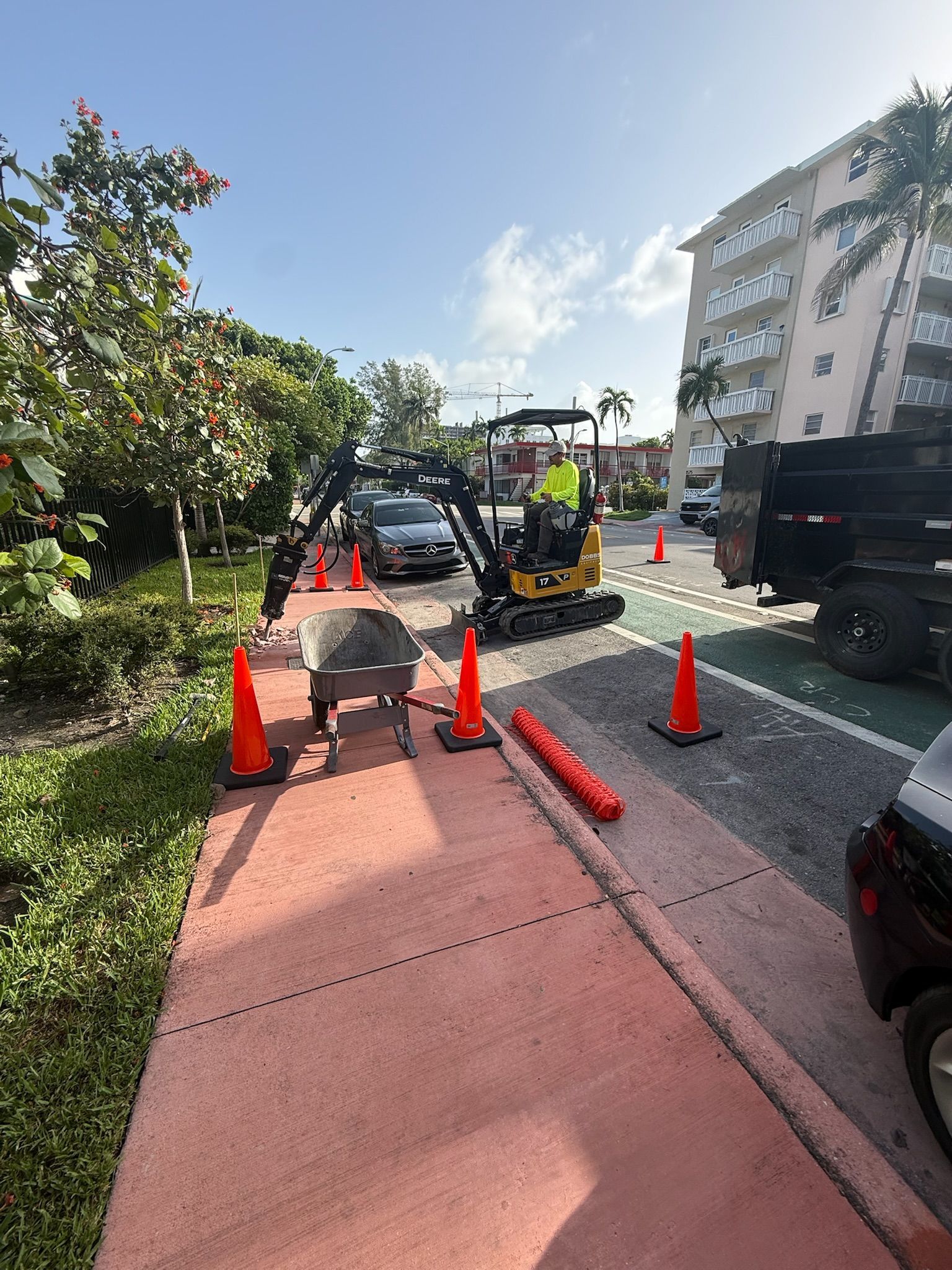 Roadwork beside a red sidewalk, with orange cones, a utility truck, and apartment buildings in sunlight