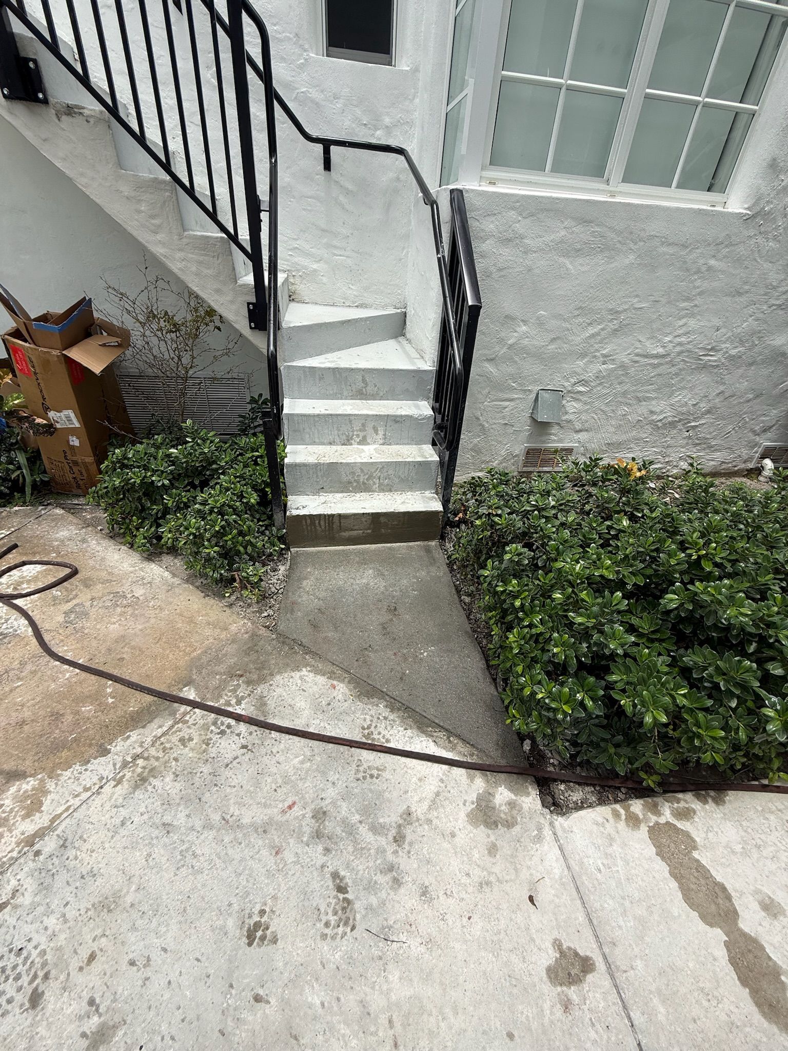 Narrow outdoor stairway beside a gray house, with black railing, concrete walkway, and green bushes.