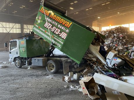 Green junk removal truck unloading trash at a waste facility.