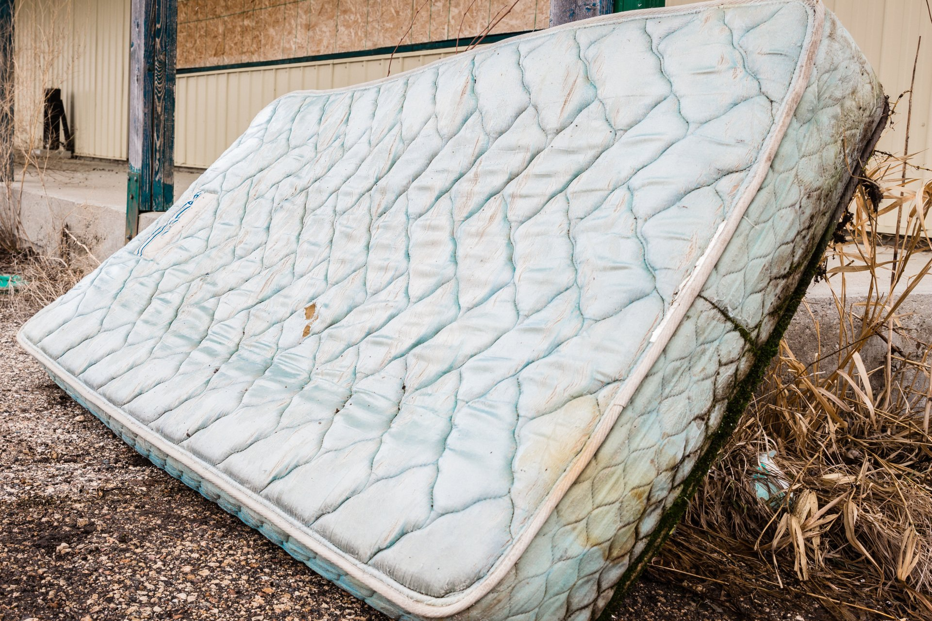 Old, stained mattress leaning against a building; outdoors, dirty.