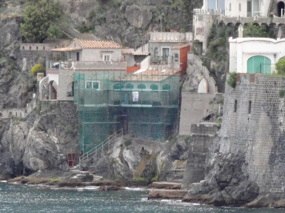 Edifici a picco sulla scogliera costiera, con reti verdi e impalcature sopra la costa rocciosa.