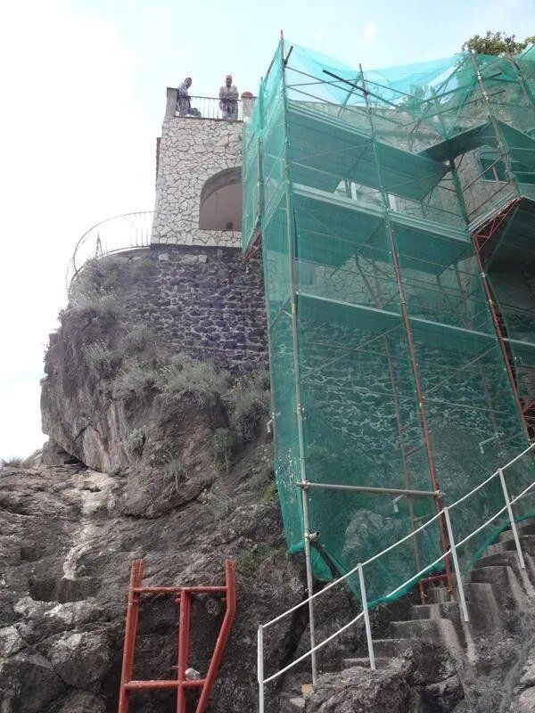 Edificio in pietra in costruzione su una scogliera rocciosa, ricoperto da una rete di impalcature verdi e da scale.
