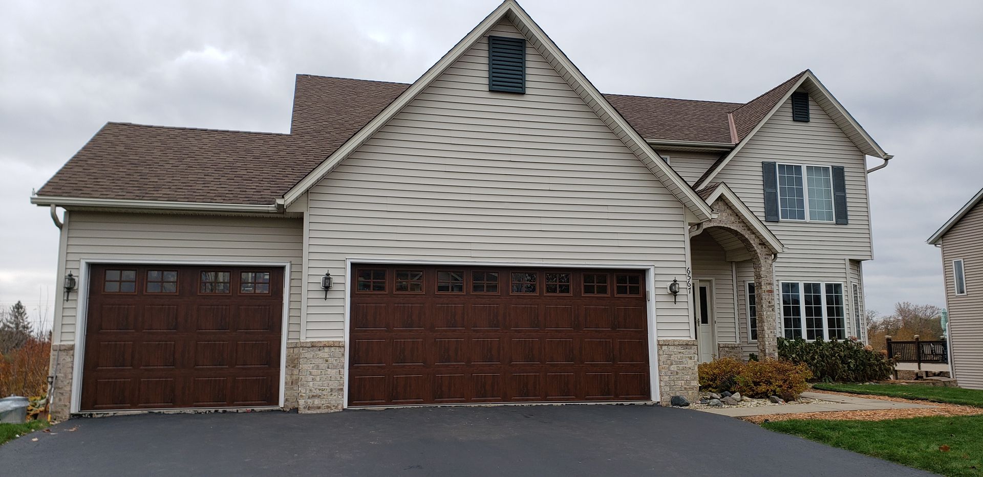 Suburban house with two garages, beige siding, and a brown roof on a cloudy day