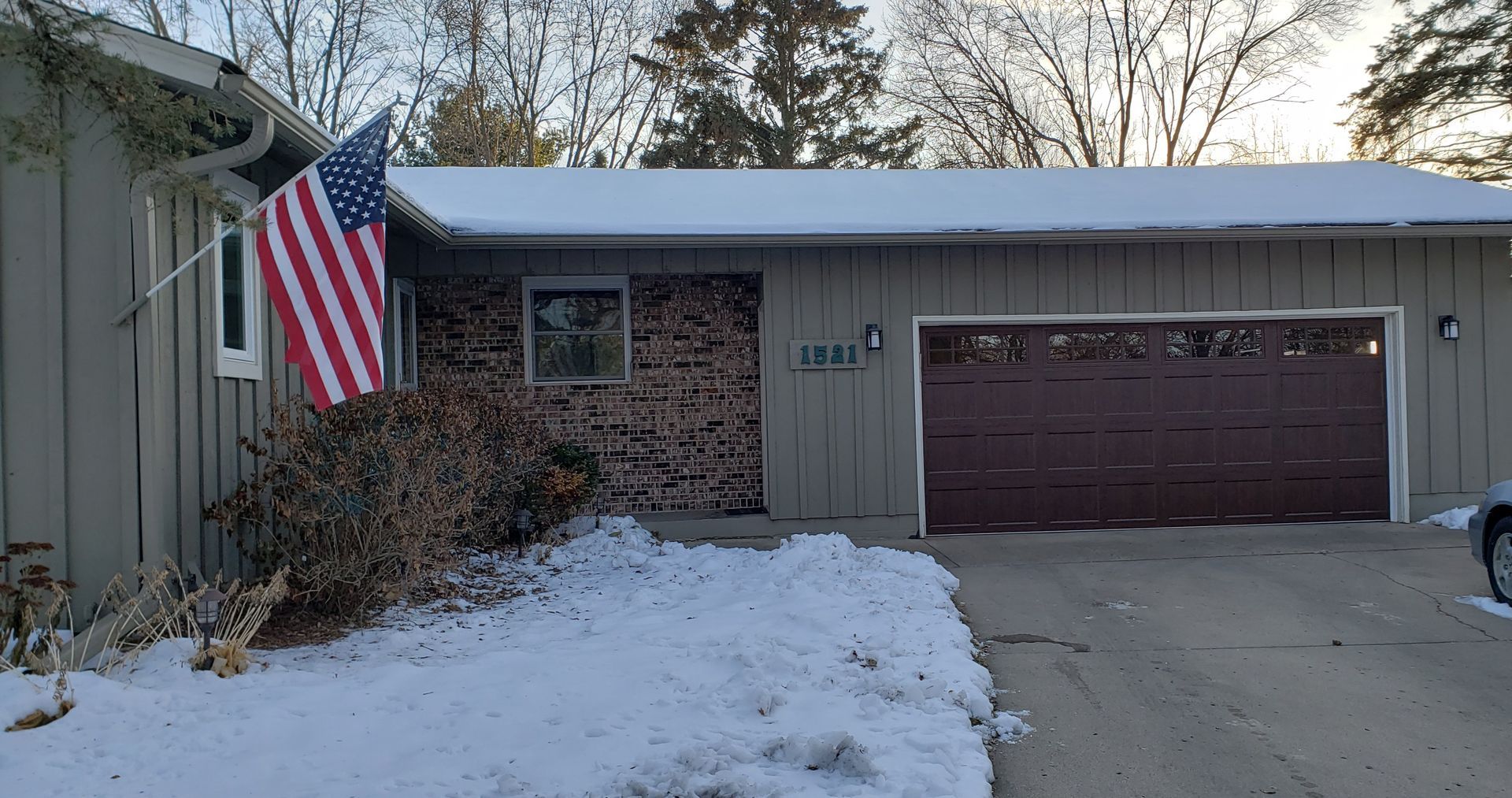 Snowy front yard with an American flag, bare trees, and a house with a garage door.