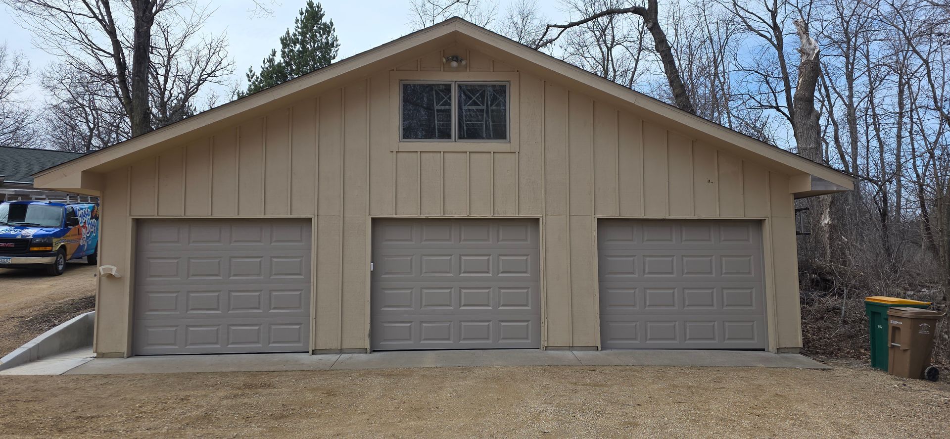 Tan garage with three closed doors and a small window above, set against bare trees
