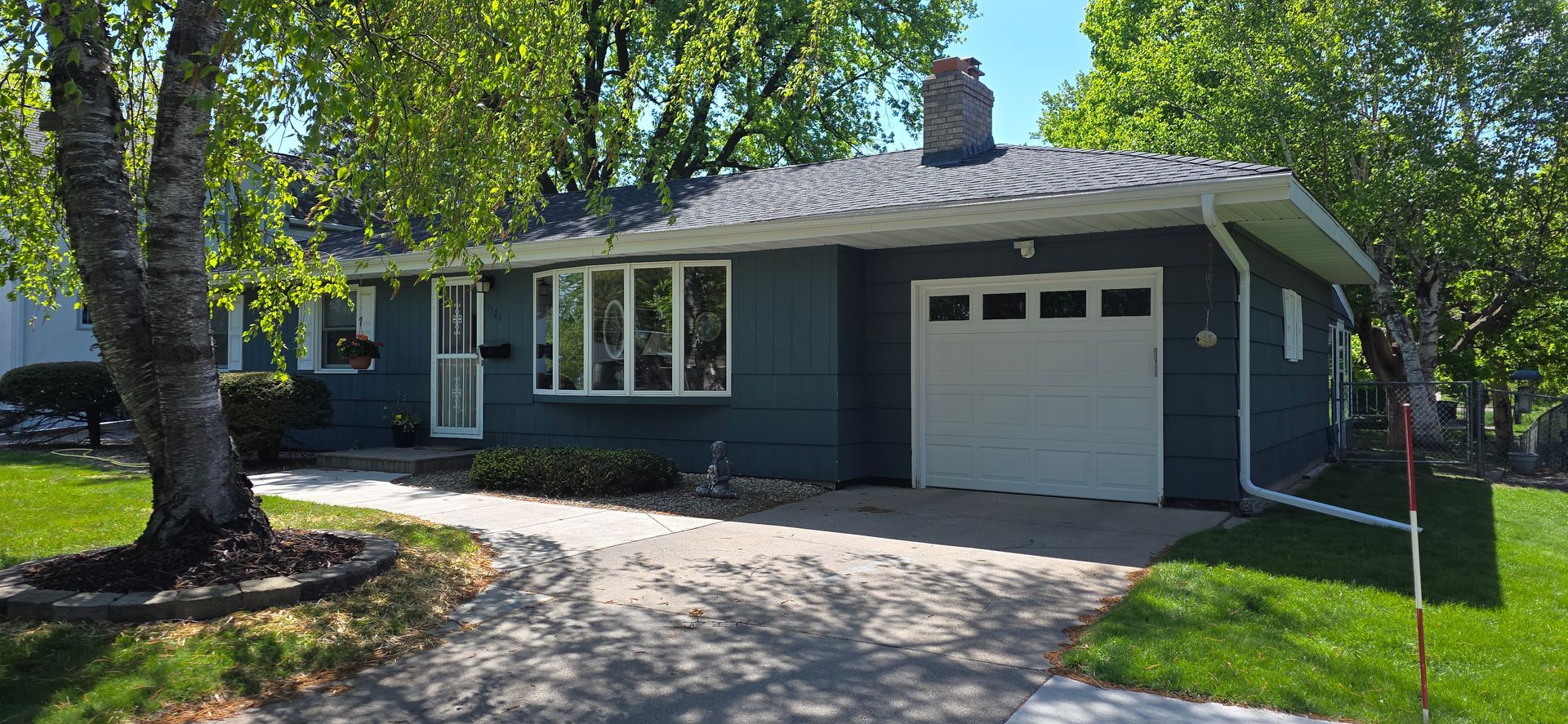 Single-story blue house with white garage, front lawn, and large tree by the driveway.