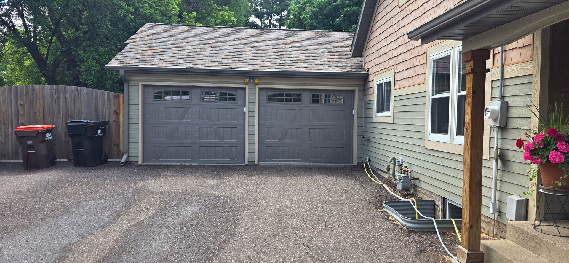 Driveway beside a beige house, with two gray garage doors, trash bins, and hanging pink flowers.