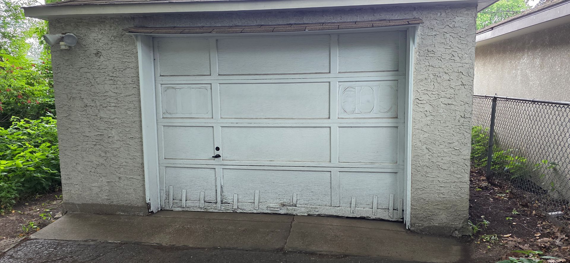Weathered white garage door set in a gray concrete outbuilding with wet pavement in front.