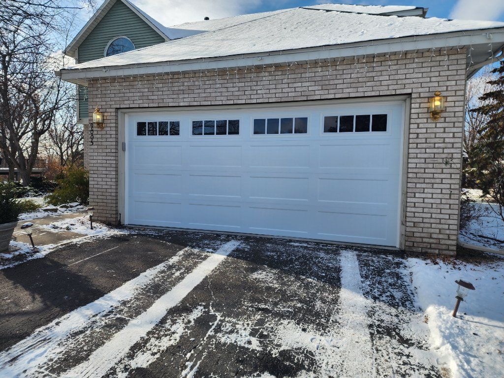 Snow-dusted white garage with a closed door and brick exterior beside a driveway in winter