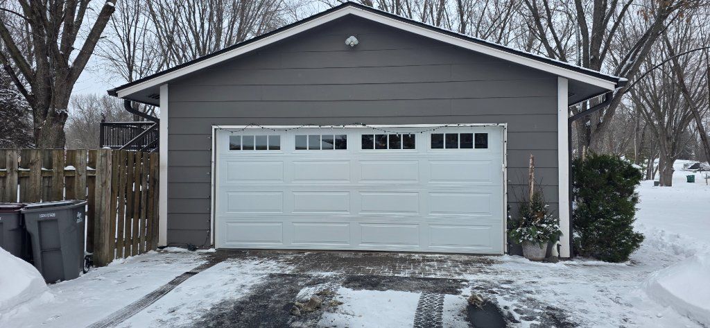 Snowy driveway leading to a detached garage with a white door and gray siding.