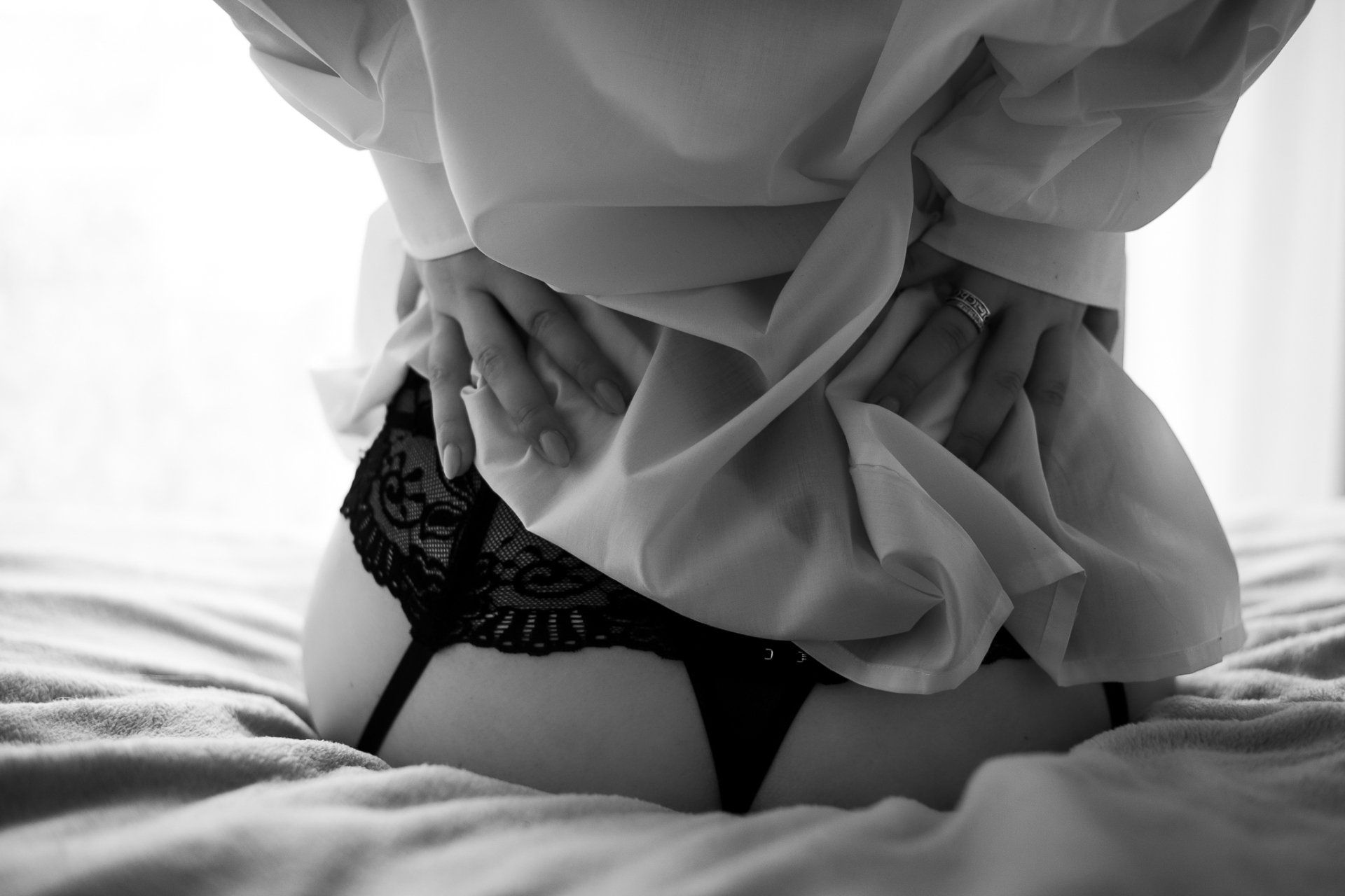 Vrouw in zwarte lingerie en witte blouse, op bed, handen op de blouse.