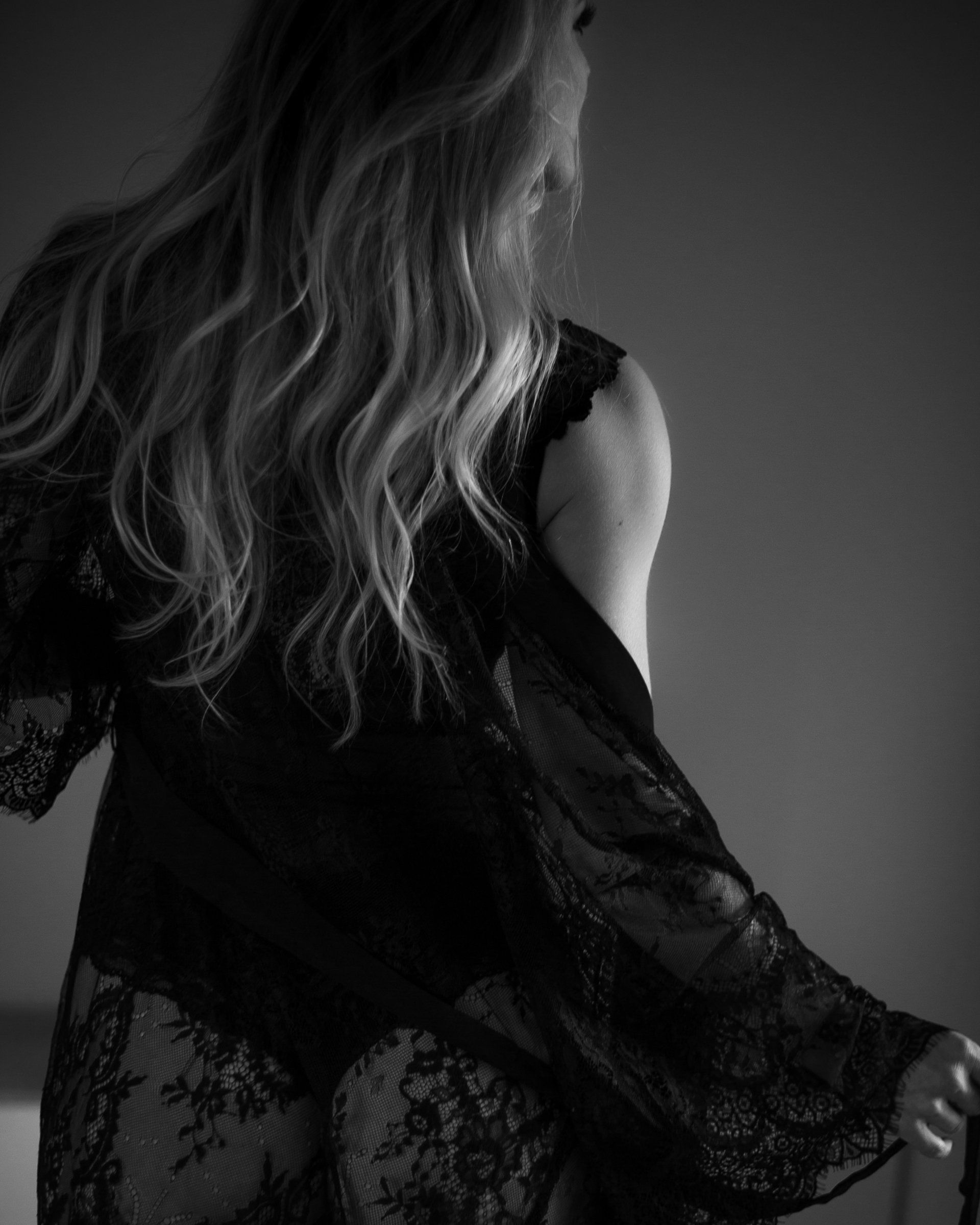 Vrouw in een zwarte kanten ochtendjas, met haar rug naar de camera gekeerd, haar haar valt in golven over haar rug.
