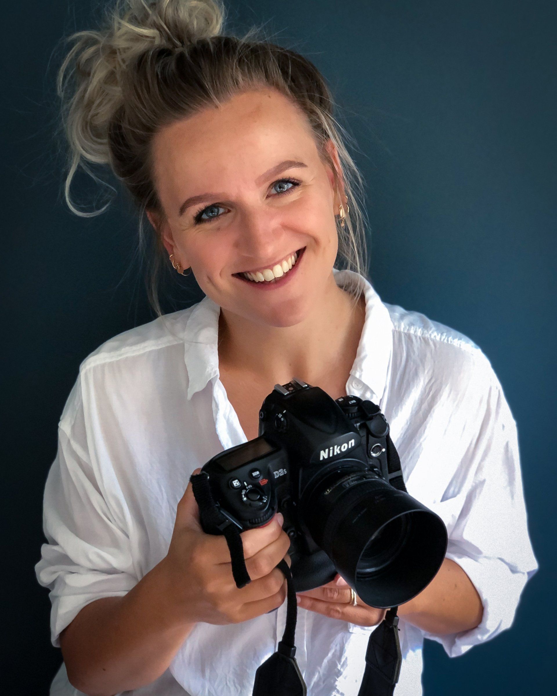 Een lachende vrouw, met een zwarte Nikon-camera met lens in haar hand, gekleed in een witte blouse, staat voor een blauwe muur.
