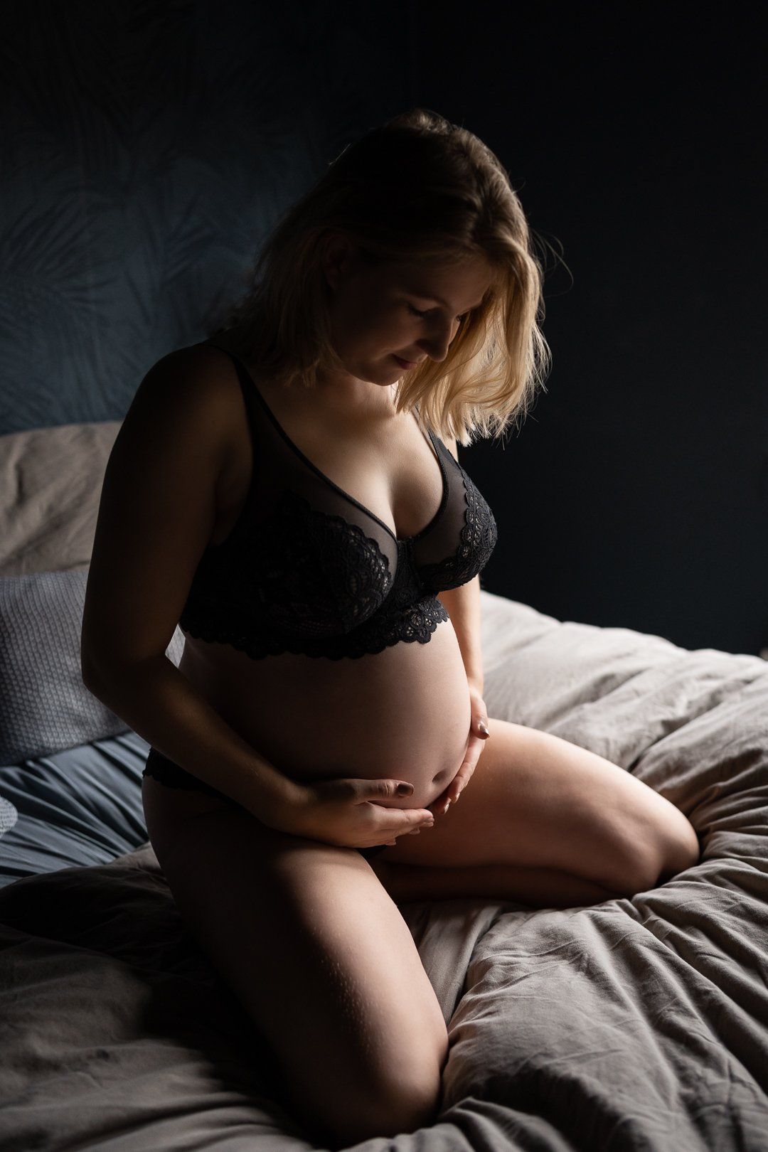 Een zwangere vrouw in zwarte lingerie zit op een bed en houdt haar buik vast.