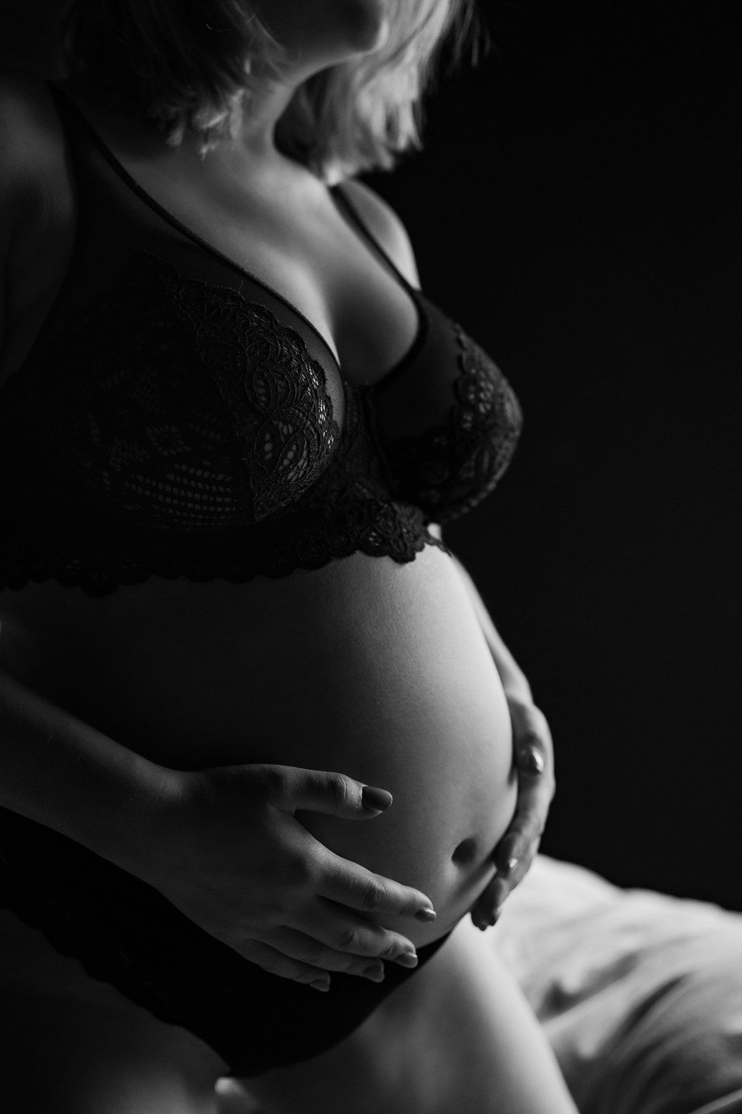 Zwangere vrouw in zwarte lingerie, die haar buik vasthoudt. Donkere achtergrond, intieme setting.