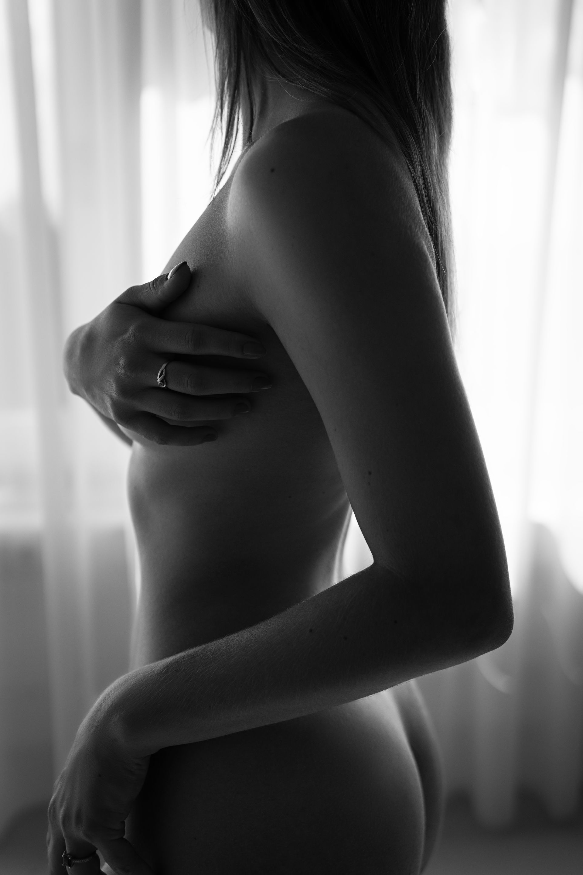 Een vrouw staat naakt, haar hand gedeeltelijk bedekt, als silhouet tegen een raam.