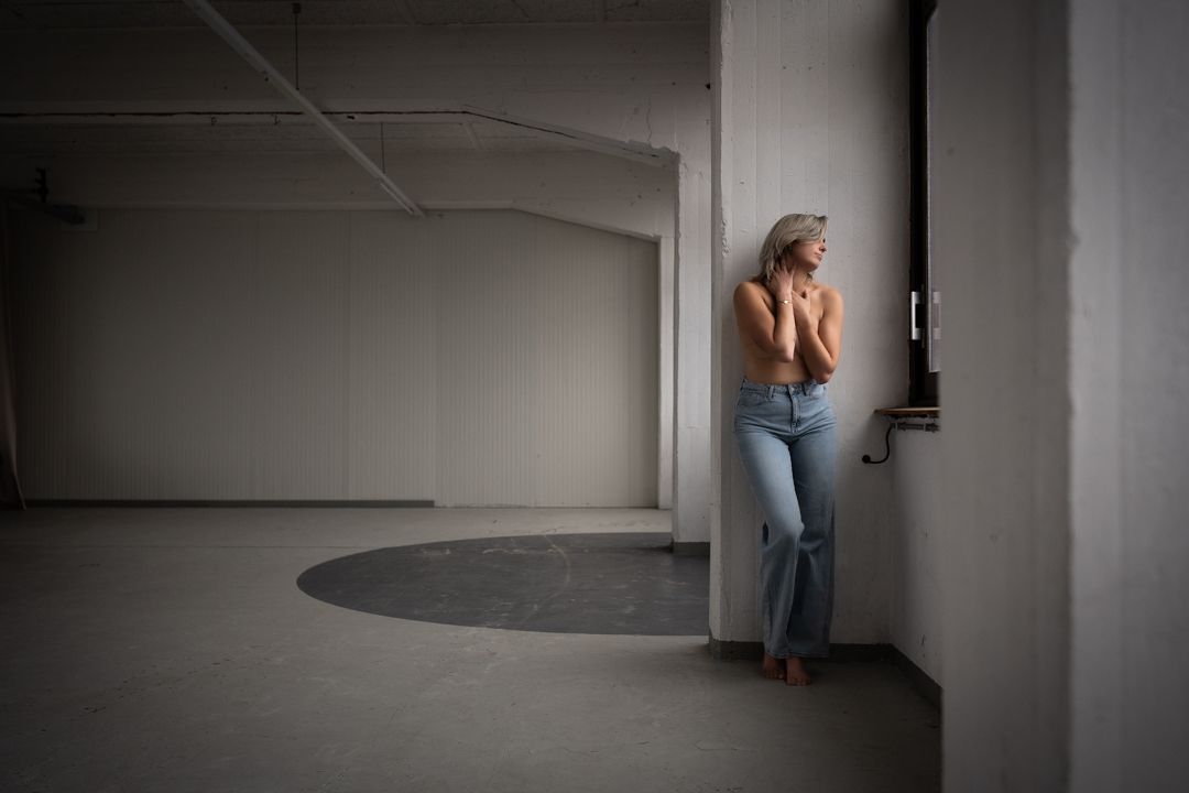 Een vrouw in spijkerbroek, met ontbloot bovenlijf, leunt tegen een raam in een grote, lege kamer en kijkt naar buiten.