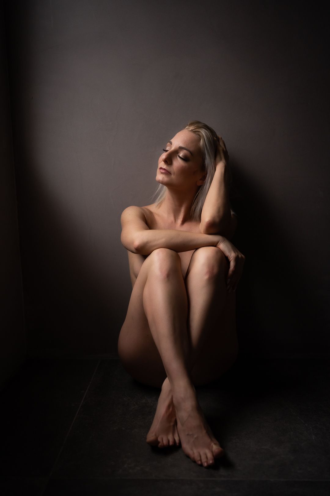Vrouw zit naakt, met haar armen over elkaar en haar ogen gesloten. Donkere omgeving, sfeervolle belichting.