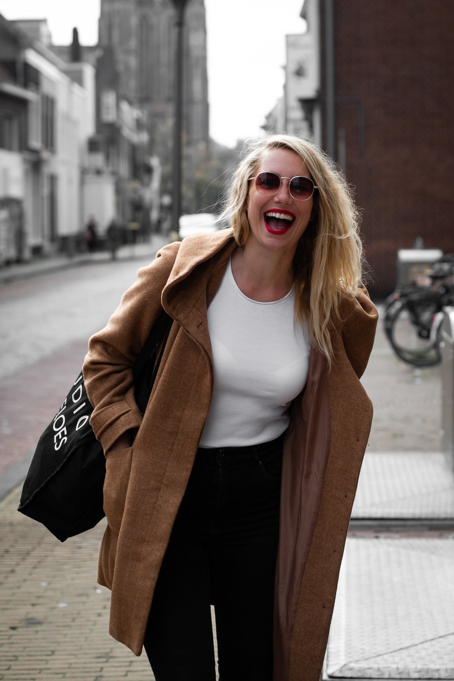 Een vrouw in een bruine jas en met een zonnebril lacht op straat.