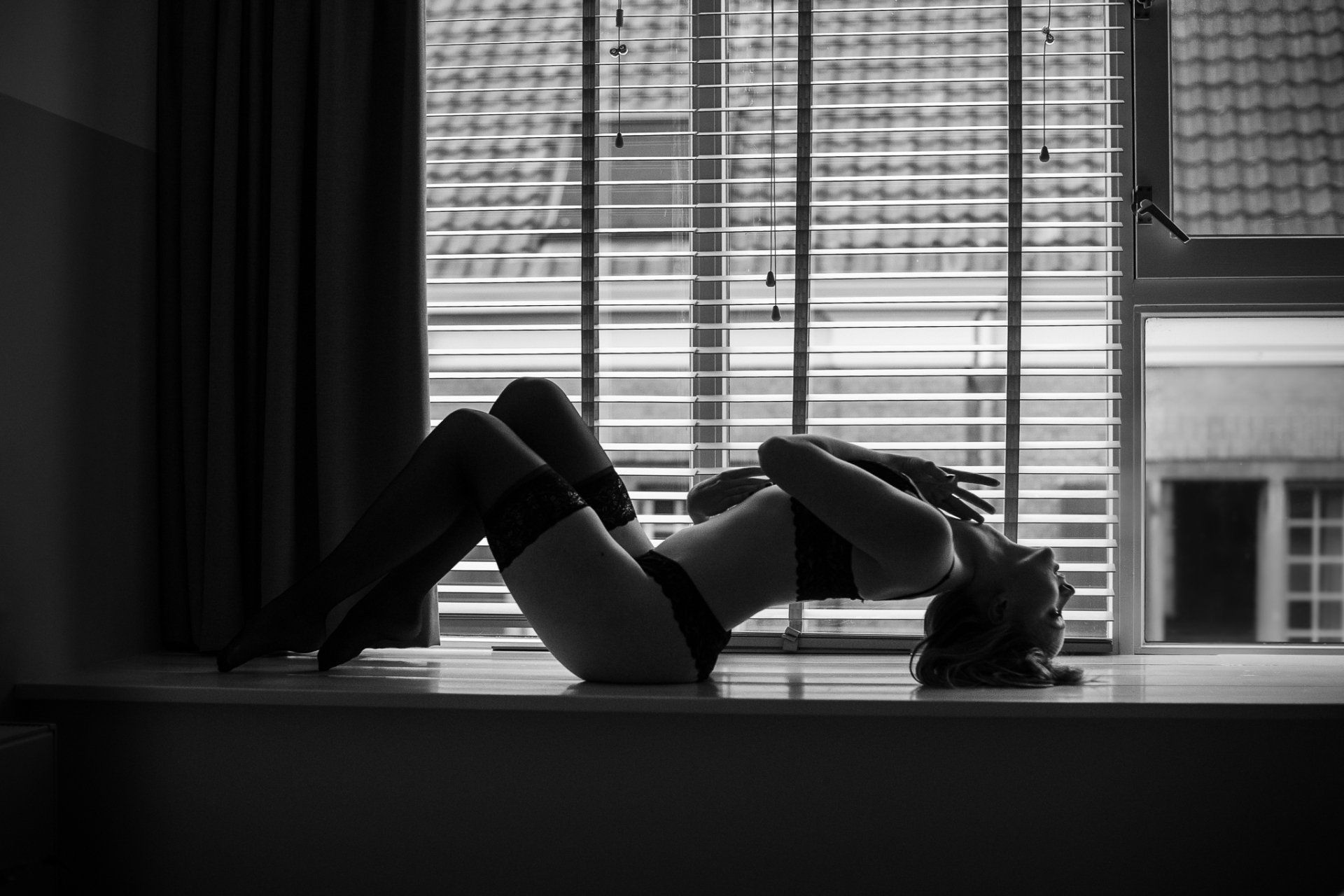 Een vrouw in lingerie buigt zich achterover op een vensterbank, haar silhouet afgetekend tegen een raam met jaloezieën.