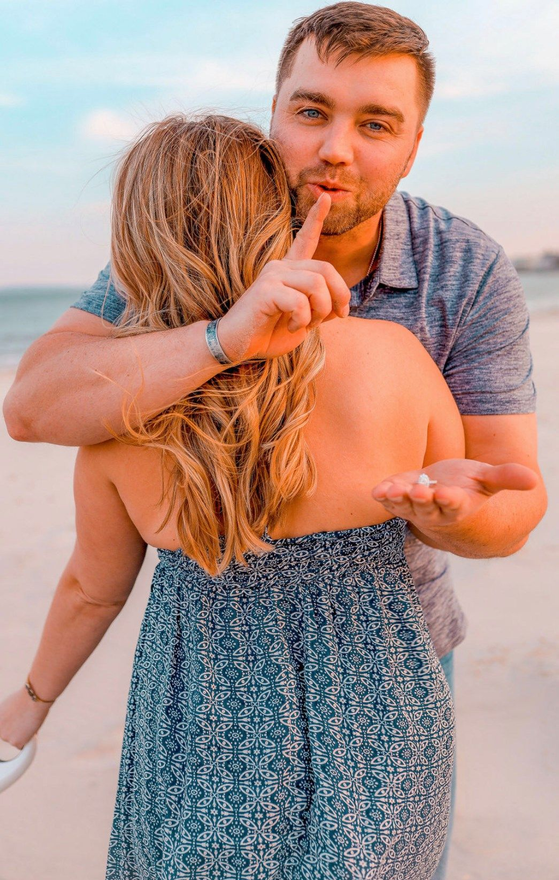 Man holding finger to lips, holding out an engagement ring as he embraces woman on beach.