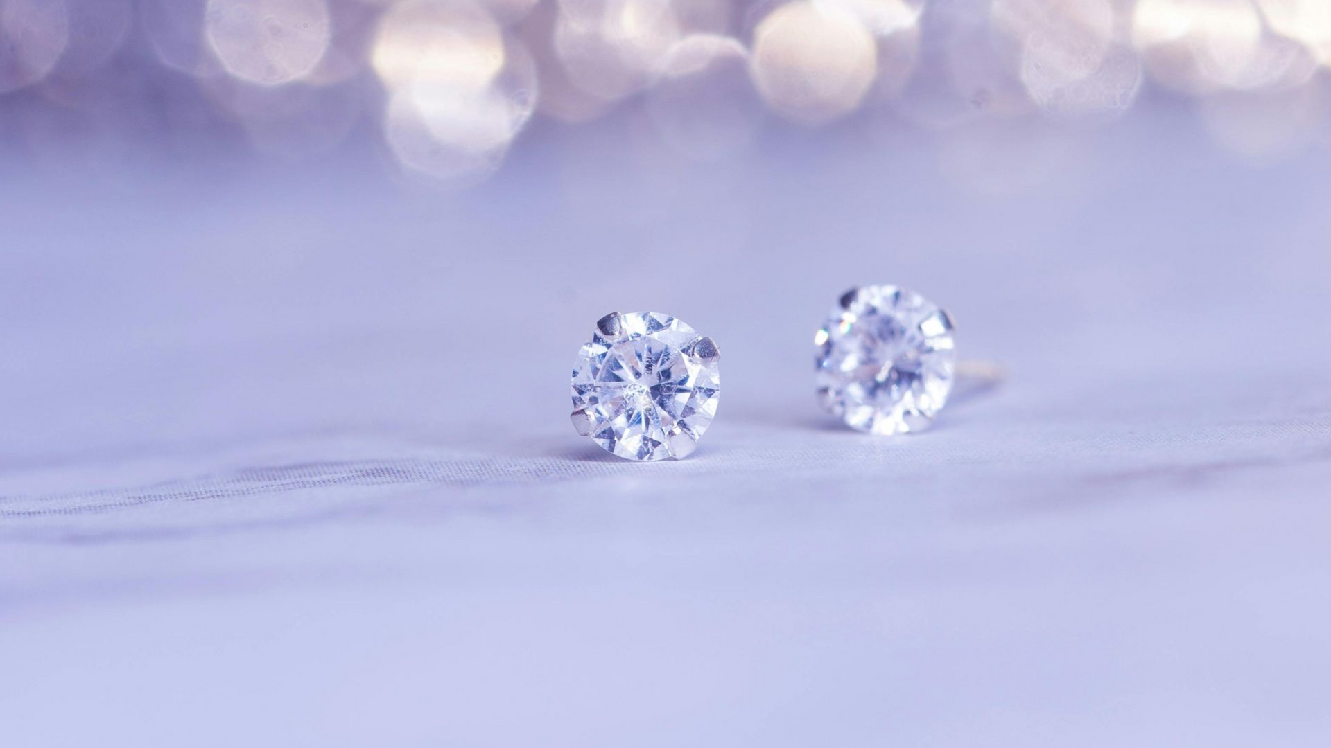 Two diamond stud earrings on a light purple surface, with bokeh background.