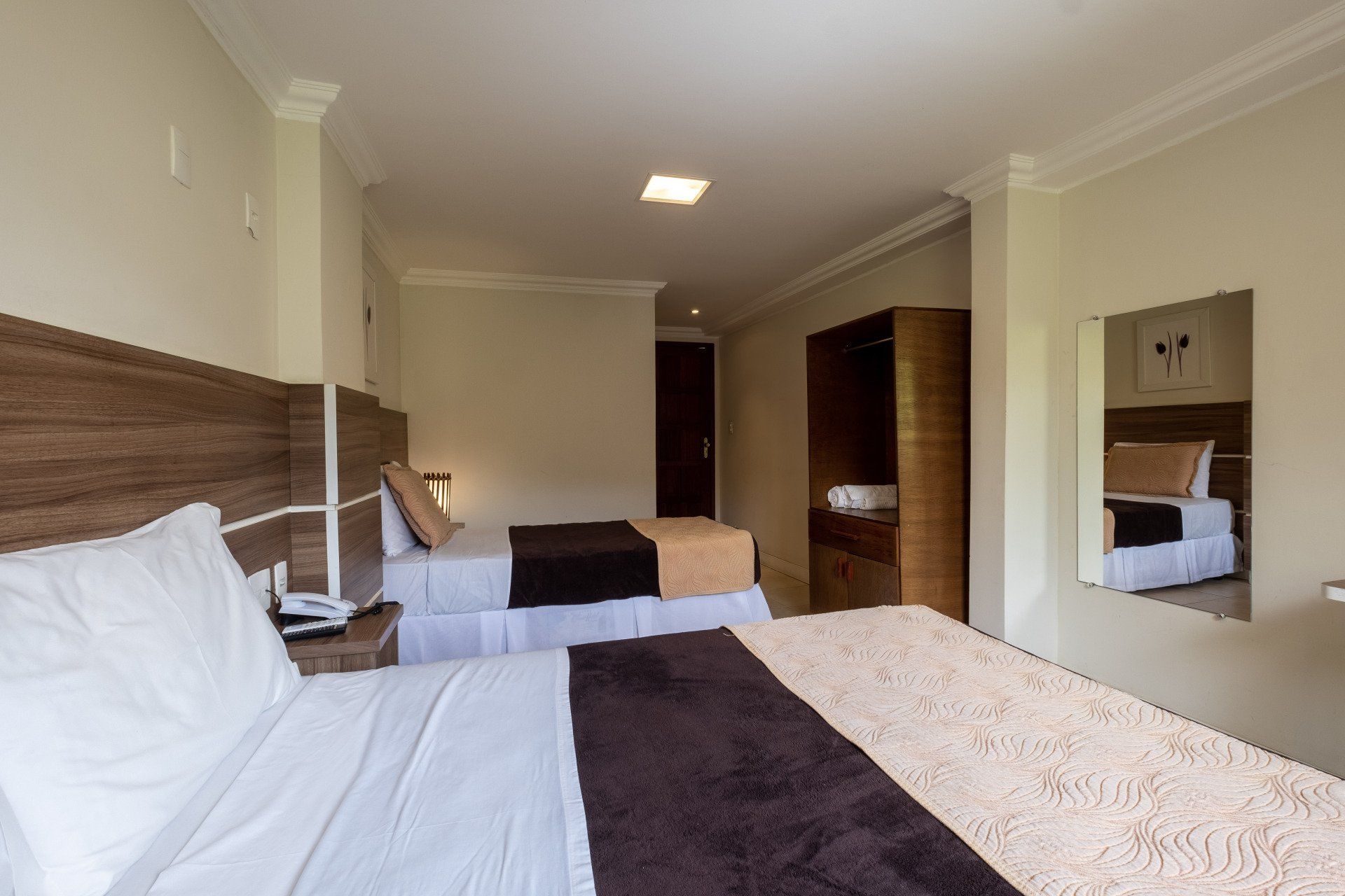 Quarto de hotel com três camas, móveis de madeira e um espelho. Decoração em tons de bege e marrom.