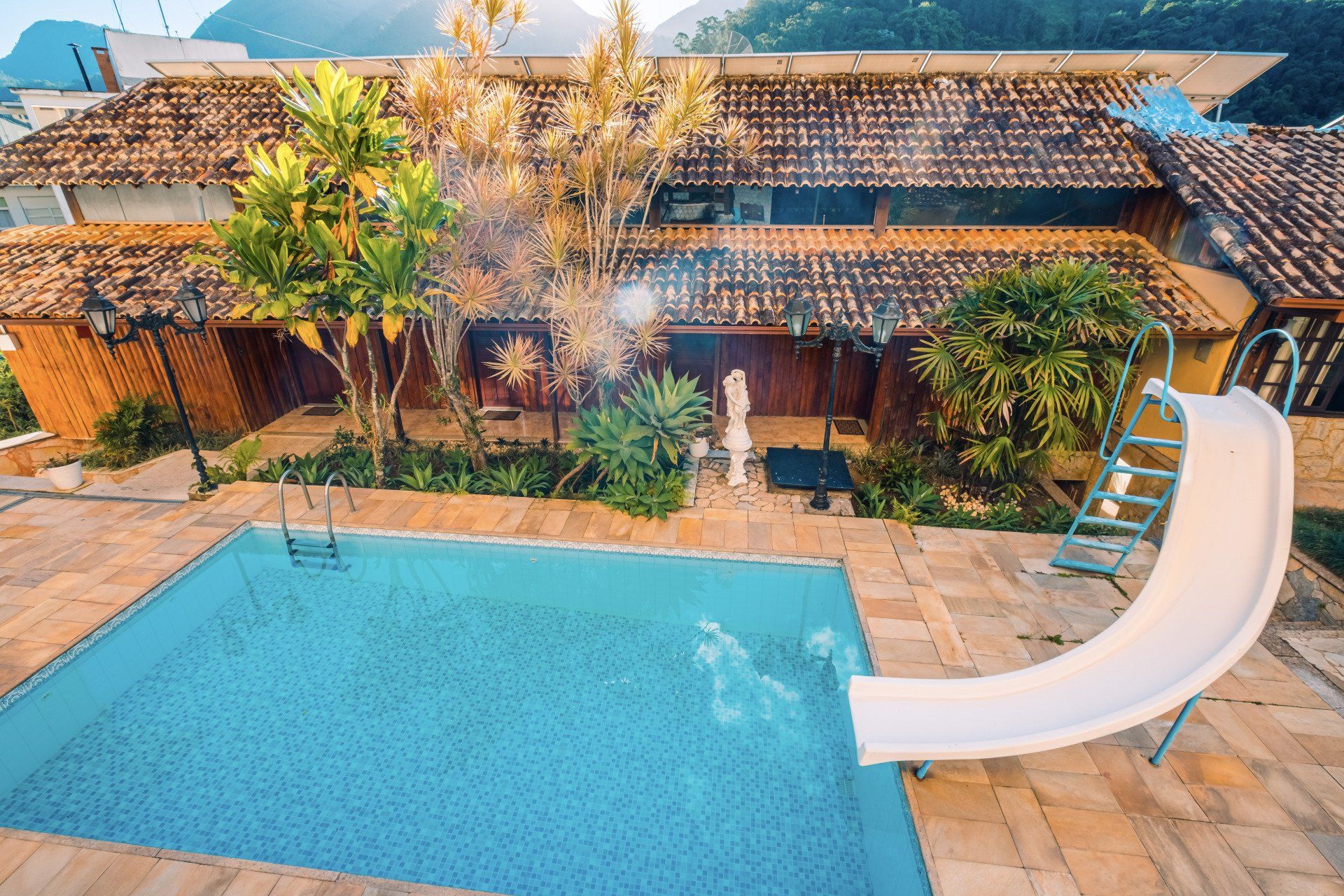 Piscina com tobogã branco, em frente a uma casa com telhado de telha. Vegetação exuberante e montanhas à vista.